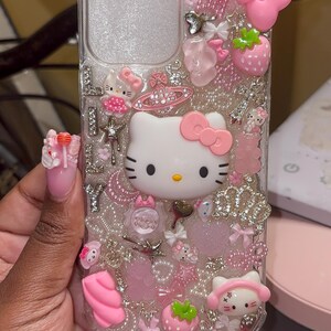 May include: A clear phone case with a pink Hello Kitty design. The case is decorated with glitter, rhinestones, and other embellishments.