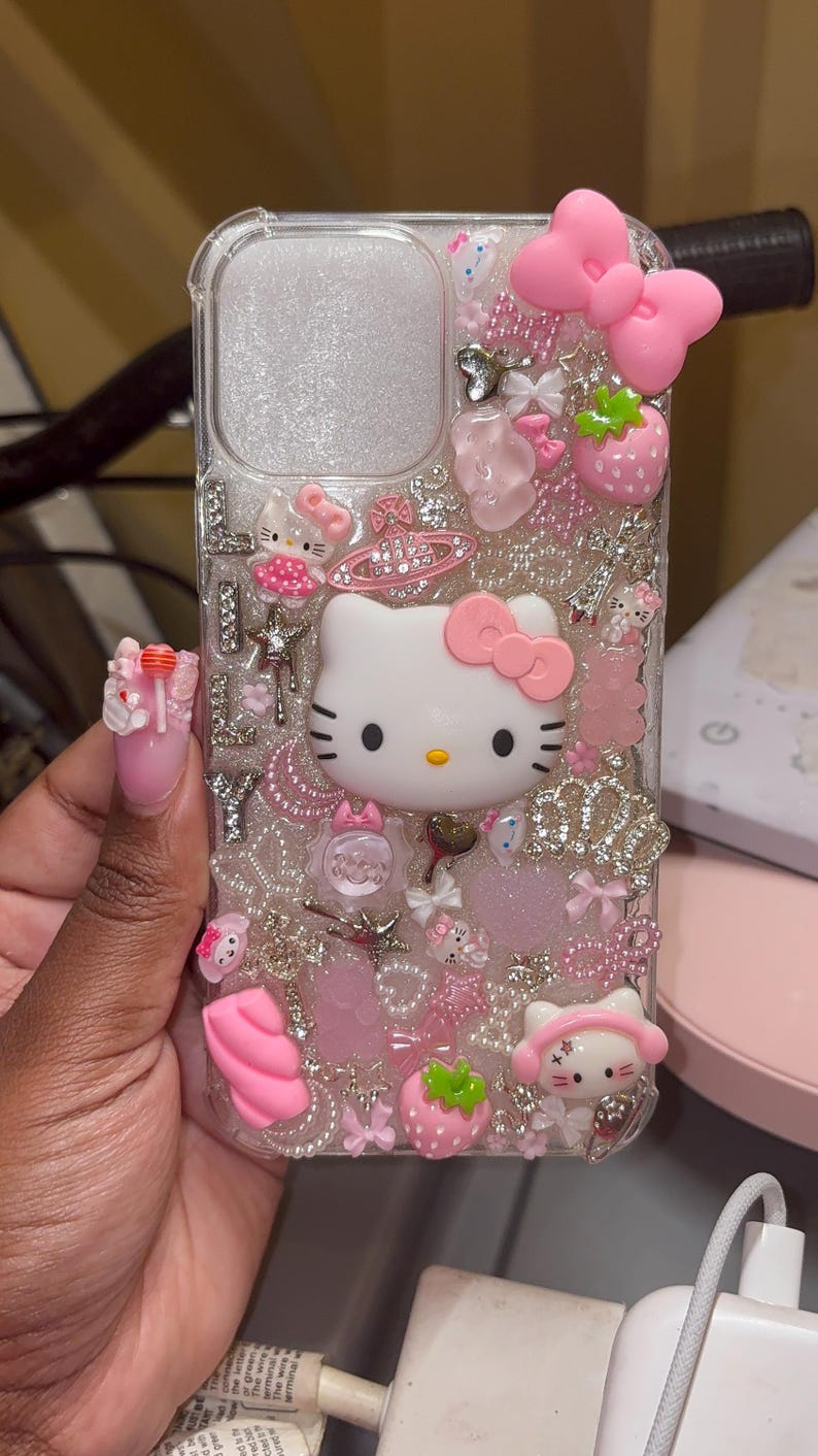 May include: A clear phone case with a pink Hello Kitty design. The case is decorated with glitter, rhinestones, and other embellishments.