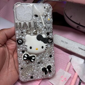 May include: A white and black Hello Kitty phone case with a pearl and rhinestone design. The case has a black bow, a black butterfly, and a black 8-ball charm.
