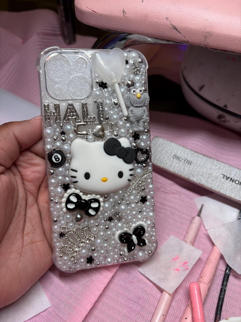 May include: A white and black Hello Kitty phone case with a pearl and rhinestone design. The case has a black bow, a black butterfly, and a black 8-ball charm.