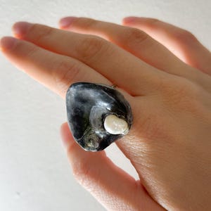 Puede incluir: Un anillo único con una piedra oscura de forma irregular y un acento blanco perlado. El diseño del anillo es orgánico y llamativo, perfecto para añadir un toque de elegancia natural. Una pieza de declaración.