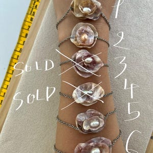 Puede incluir: Seis pulseras con colgantes de concha en forma de flor y centros de perlas se muestran en una muñeca. Las pulseras están conectadas por una cadena plateada. La palabra "SOLD" está escrita en dos de las pulseras. Una cinta métrica amarilla es visible en el fondo.