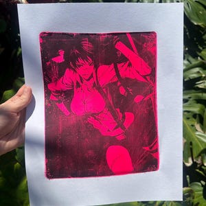 以下が含まれることがあります： 白い紙に描かれた、ピンクと黒のアニメ風キャラクターのプリント。このアートワークは、暗い髪と決意に満ちた表情を持つ人物を、大胆な線と鮮やかなピンクの色合いで表現しています。プリントは緑の葉の背景に保持されています。
