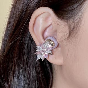 Puede incluir: Un brazalete de oreja de plata y cristal con un diseño floral. El brazalete presenta un grupo de cristales transparentes y rosas dispuestos en forma de flor, con una base de metal dorado y un auricular de color lavanda.