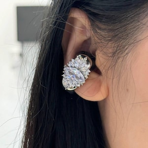 Puede incluir: Primer plano de un pendiente de plata y cristal transparente. El pendiente presenta un gran cristal de forma ovalada rodeado de cristales más pequeños en forma de hoja. El pendiente está sobre una base negra y se lleva en la oreja.