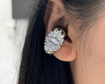 May include: Close-up of a silver and clear crystal earring. The earring features a large, oval-shaped crystal surrounded by smaller, leaf-shaped crystals. The earring is on a black base and is worn on the ear.