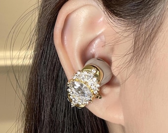 May include: A gold-coloured earbud featuring a large, oval-shaped clear crystal at its centre, encircled by smaller clear crystals. The earbud is positioned against a light-coloured ear. The design is ornate and elegant, resembling a piece of jewellery.
