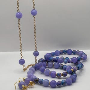 May include: A jewelry set consisting of a long gold-tone chain necklace with lavender beads, three beaded bracelets featuring a mix of solid lavender and blue-purple patterned beads, and a pair of matching gold-tone drop earrings with lavender beads.