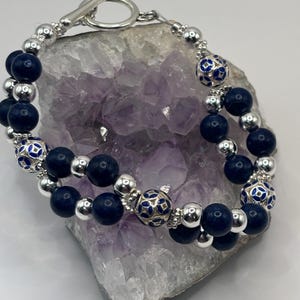 Op de afbeelding: Een zilveren armband met marineblauwe en zilveren kralen. De armband heeft decoratieve blauwe en zilveren kralen met een ingewikkeld ontwerp. De tuimelsluiting is zilver. De armband wordt gepresenteerd op een paars kristal.