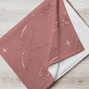 Könnte beinhalten: Eine staubrosa Decke mit weißer Unterseite. Die Decke zeigt ein sich wiederholendes Muster aus einem Drachen, Rosen, Kronen, Büchern und Sternen. Die Decke ist gefaltet und zeigt die weiße Unterseite.