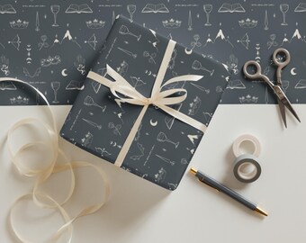 May include: A wrapped gift with a cream-colored ribbon, scissors, tape, and a pen on a white surface. The gift wrap is dark gray with a white pattern of moons, crowns, swords, and other mystical symbols. The background is a matching pattern.