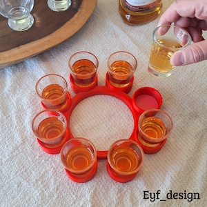Op de afbeelding: Een rode shotglashouder met acht shotglaasjes gevuld met amberkleurige vloeistof. Eén shotglaasje wordt vastgehouden. De houder heeft een cirkelvormig ontwerp met individuele sleuven. De tekst "Eyf_design" is zichtbaar.