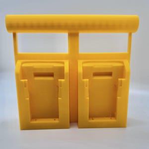 Puede incluir: Un soporte de pared de plástico amarillo con dos ranuras para guardar baterías. El soporte tiene una barra horizontal en la parte superior.