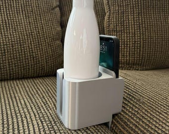 May include: A white water bottle with a silver lid is in a silver 3D-printed cup holder. A black smartphone shows 8:51. The cup holder is attached to a brown sofa. The product is from TWO 3D PRINTERS.
