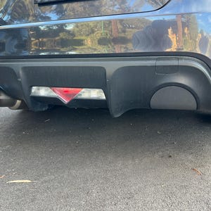 May include: The rear bumper of a dark gray vehicle features a black diffuser with a central triangular red and white light assembly. A single circular exhaust pipe with a blue-tinted metallic tip is visible on the left side of the bumper assembly.