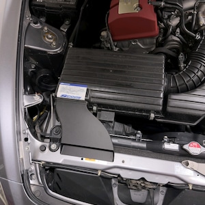 May include: A black plastic air intake box with a Spoon Sports sticker featuring blue and white text, installed in a car engine bay next to a red valve cover with gold Honda VTEC lettering and various black rubber hoses and metal components.