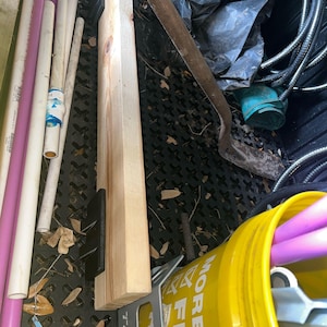 May include: A collection of construction and gardening supplies including several purple and white PVC pipes, a rectangular wooden board with black plastic brackets, a metal shovel, a black plastic bag, and a yellow bucket with white text.