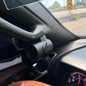 Op de afbeelding: Een matzwarte cilindrische meterhouder bevestigd aan de A-stijl van een voertuiginterieur. De houder is voorzien van het woord Cummins in reliëf aan de zijkant. Het interieur bevat een donker dashboard, een stuurwiel en een gedeeltelijke meter.