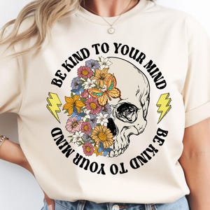 Op de afbeelding: Crèmekleurig T-shirt met een schedelontwerp met kleurrijke bloemen en een vlinder. De tekst "BE KIND TO YOUR MIND" is rond de schedel gedrukt. Twee gele bliksemschichten flankeren de schedel.