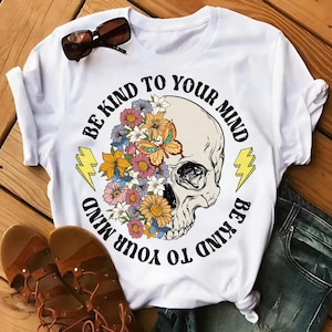May include: White t-shirt with a skull design. The skull is adorned with colorful flowers and a butterfly, with the text "BE KIND TO YOUR MIND" in a circular pattern. Two yellow lightning bolts flank the skull.