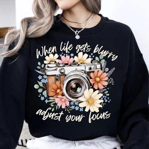 May include: Black sweatshirt featuring a vintage camera design surrounded by colorful flowers. The text reads "When life gets blurry, adjust your focus."