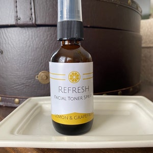 May include: A brown glass bottle of Refresh Facial Toner Spray with a white label featuring a lemon graphic. The label reads "Lemon & Grapefruit." The bottle has a black spray top and a clear plastic cap. The bottle is on a white rectangular tray.