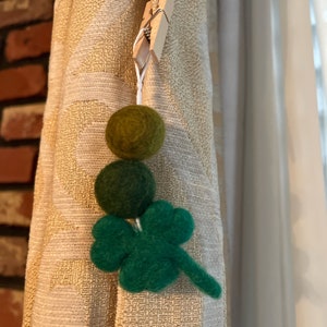 May include: A green felt shamrock with three leaves hanging from a white string with two green felt balls. The string is attached to a wooden clothespin.