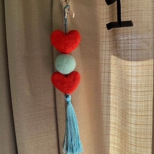 May include: A red felt heart with a blue tassel hanging from a wooden clothespin. The heart is attached to a smaller green felt ball and another red felt heart.