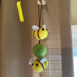 May include: A felt bee garland with two yellow and black bees and a green felt ball hanging from a brown string.