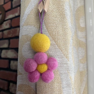 May include: A yellow felt ball with a pink felt flower attached to it. The flower has a yellow felt center. The ball and flower are hanging from a purple string.