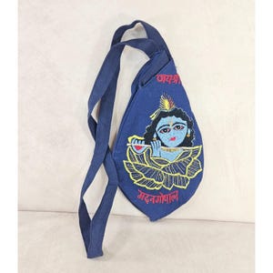 May include: A teardrop-shaped navy blue fabric bag with a long shoulder strap. The front features an embroidered illustration of a blue-skinned figure playing a flute, surrounded by yellow lotus petals, with red text in Devanagari script above and below.
