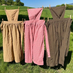 May include: Three aprons hang on a clothesline in a sunny outdoor setting. The aprons are in shades of tan, pink with a white floral pattern, and brown. The aprons are made of fabric and are hanging in front of a green field.