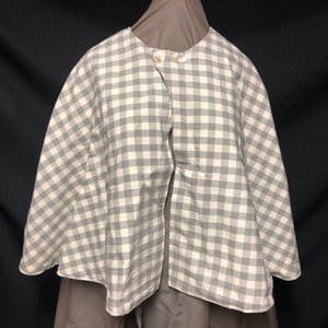 Op de afbeelding: Een grijs en wit geruite cape met twee houten knopen. De cape is aan de voorkant open en heeft een ronde halslijn. De stof lijkt katoen of een vergelijkbaar materiaal te zijn.