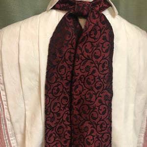 Puede incluir: Un ascot burdeos con un estampado floral negro está drapeado sobre una camisa blanca con cuello. El ascot está hecho de una tela texturizada y está atado con un lazo en el cuello. La camisa tiene pliegues verticales.