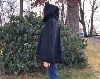 May include: A black hooded cloak with a cape-like design. The cloak is made of a smooth, dark fabric and features a hood that covers the head. The person is wearing blue jeans. The background includes green bushes and trees.