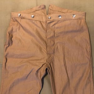 Puede incluir: Pantalones de color beige con cintura alta y cierre de botones. Los pantalones tienen un diseño de pierna recta y presentan botones plateados. La tela parece ser de algodón o mezcla de peso medio.