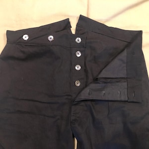 Puede incluir: Pantalones negros de cintura alta con cierre de botones. Los pantalones tienen una cintura ancha con dos botones a cada lado y una serie de botones en la parte delantera. La tela parece ser de un color oscuro y liso.