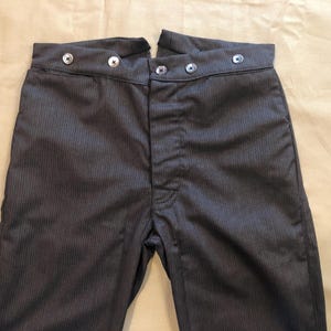 Puede incluir: Primer plano de un pantalón gris carbón con finas rayas verticales. El pantalón de cintura alta presenta un cierre de botones y seis botones decorativos en la cintura. La tela tiene un brillo sutil, lo que sugiere un estilo formal o vintage.