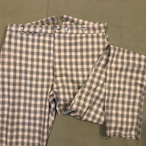 Puede incluir: Pantalones a cuadros color crema con un patrón de cuadrícula negro y marrón. Los pantalones tienen una cintura alta con detalles de botones plateados. La tela está doblada, mostrando el interior. Los pantalones están sobre una superficie verde.