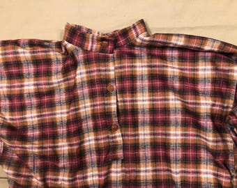 May include: A close-up of a plaid shirt in shades of burgundy, brown, and white. The shirt features a mandarin collar and three brown buttons down the front. The fabric has a soft, textured appearance.
