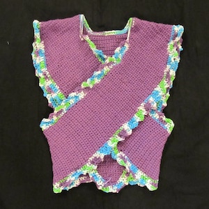 May include: A handmade, purple crochet vest with a wrap-around design. The edges are trimmed with a scalloped border in white, green, and blue. The vest is displayed on a black background, showcasing the intricate stitch work and color details.