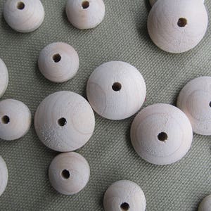 May include: A collection of unfinished wooden beads in various sizes, each with a central hole. The beads are light-colored and appear to be made of natural wood, with visible grain patterns. They are scattered on a textured, neutral-toned surface.