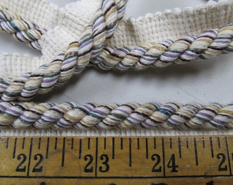 May include: Close-up of decorative rope trim with a twisted design. The rope features a cream-colored base with stripes of purple, green, gold, and brown. A wooden ruler with inch markings is visible at the bottom, measuring from 22 to 25 inches.