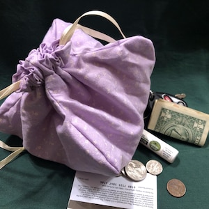 May include: A lavender drawstring bag with a cream-colored ribbon closure. The bag is filled with items including a dollar bill, coins, keys, and a small tube. The bag has a floral pattern.