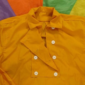 May include: A close-up of a mustard yellow shirt with a double-breasted front and white buttons. The shirt has a classic collar and is made of a smooth fabric. The background is a colorful patchwork of orange, green, purple, and yellow.