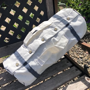 May include: A white shirt-shaped bag with blue stripes and button details. The bag has a cylindrical shape and is made of a light-colored fabric. The buttons are a light brown color. The bag is resting on a wooden bench.