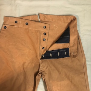 Puede incluir: Pantalones de trabajo color canela con bragueta abotonada y botones negros. Los pantalones tienen cintura alta y corte holgado. El interior de la cinturilla es azul marino. Los pantalones son de material resistente.