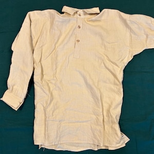 May include: A light beige long-sleeved shirt with a collar and three wooden buttons down the front. The shirt has a relaxed fit and is made of a textured fabric. The sleeves have buttoned cuffs. The shirt is laying on a dark green surface.