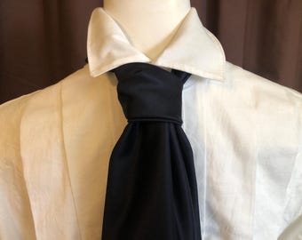 May include: A white dress shirt with a classic collar and a dark navy ascot tie. The tie is knotted at the neck and drapes down the front of the shirt. The shirt has vertical pleats.