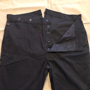 Puede incluir: Pantalones azul marino oscuro con cintura alta y cierre de botones. Los pantalones tienen un diseño clásico con múltiples botones en la parte delantera y lateral. La tela parece ser un material duradero.